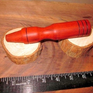 Puede incluir: Un reclamo de pato de madera de color marrón rojizo apoyado sobre dos pequeñas rodajas de madera. El reclamo tiene una boquilla cónica y un cuerpo más ancho con tres ranuras oscuras. Una regla negra está en primer plano.