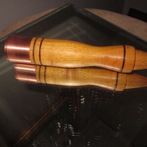 Puede incluir: Dos herramientas de madera con extremos de cobre y líneas negras. Las herramientas tienen un mango redondeado y un extremo cónico. Las herramientas están sobre una superficie reflectante.