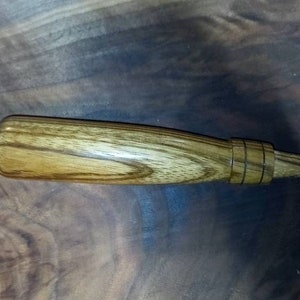 May include: A wooden duck call with a light brown and dark brown wood grain pattern. The call has a rounded handle and a tapered mouthpiece with two dark bands near the end. The duck call is a hunting accessory.