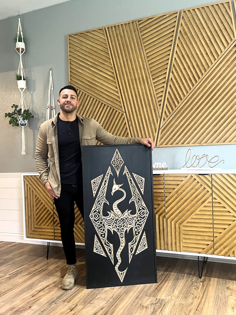 K&ouml;nnte beinhalten: Ein schwarzes Wandkunstst&uuml;ck aus Holz mit einer detaillierten Schnitzerei eines Drachen in einem keltischen Knotendesign. Der Drache ist in einer hellen Holzfarbe geschnitzt und vor einem schwarzen Hintergrund platziert.