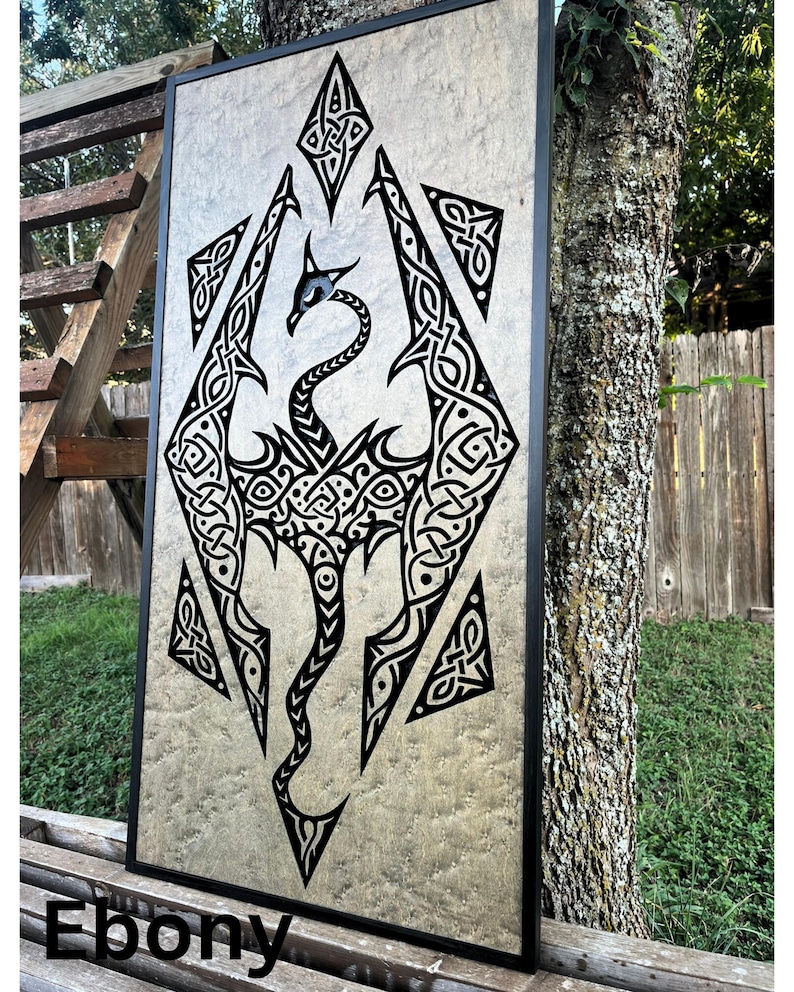 K&ouml;nnte beinhalten: Ein schwarzes und wei&szlig;es Drachen-Design mit komplizierten Knotenarbeiten, gerahmt in schwarzem Holz. Der Hintergrund ist in Metallic-Gold gehalten. Das Wort "Ebony" ist unter dem Rahmen gedruckt.