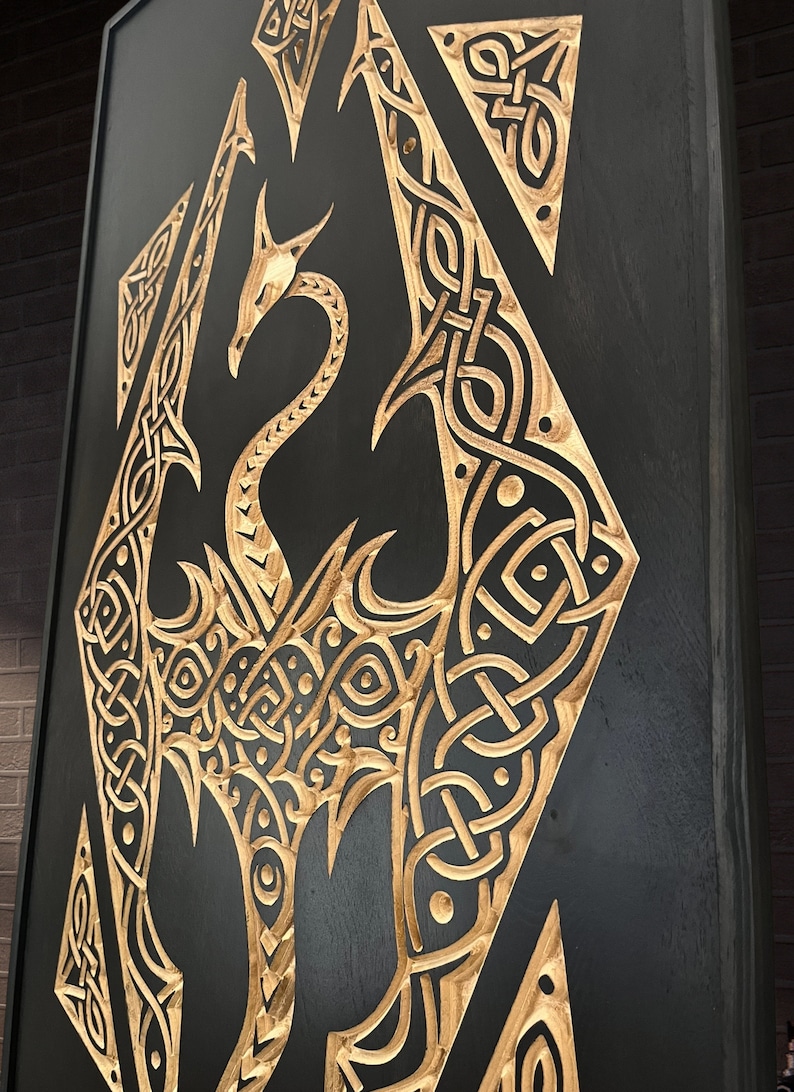 K&ouml;nnte beinhalten: Eine schwarze Holztafel mit einem goldenen Drachenmotiv, das hineingeschnitzt ist. Der Drache ist in einem keltischen Knotenmuster.