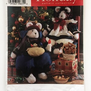 May include: Simplicity Holiday pattern collection sewing pattern for two plush teddy bears. One bear is wearing a blue and red outfit with a brown hat and the other is wearing a white dress with a red bow. The pattern is for one size and is called "Holiday".