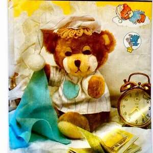 Puede incluir: Un patrón Simplicity crafts/ouvrages para hacer un oso de peluche con pijama. El patrón incluye una manta azul, un libro y un despertador. El patrón se titula "Teddy Beddy Bear" y es de 1986.