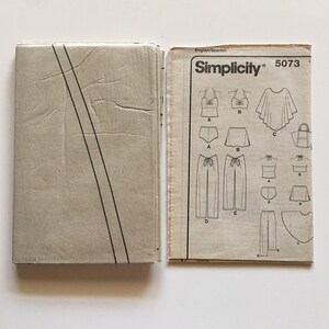 Puede incluir: Un patr&oacute;n de costura Simplicity vintage, modelo 5073, con ilustraciones de varias prendas de vestir, incluyendo tops, shorts, pantalones y un bolso tote. El patr&oacute;n est&aacute; en ingl&eacute;s y espa&ntilde;ol, con una combinaci&oacute;n de colores beige y blanco.