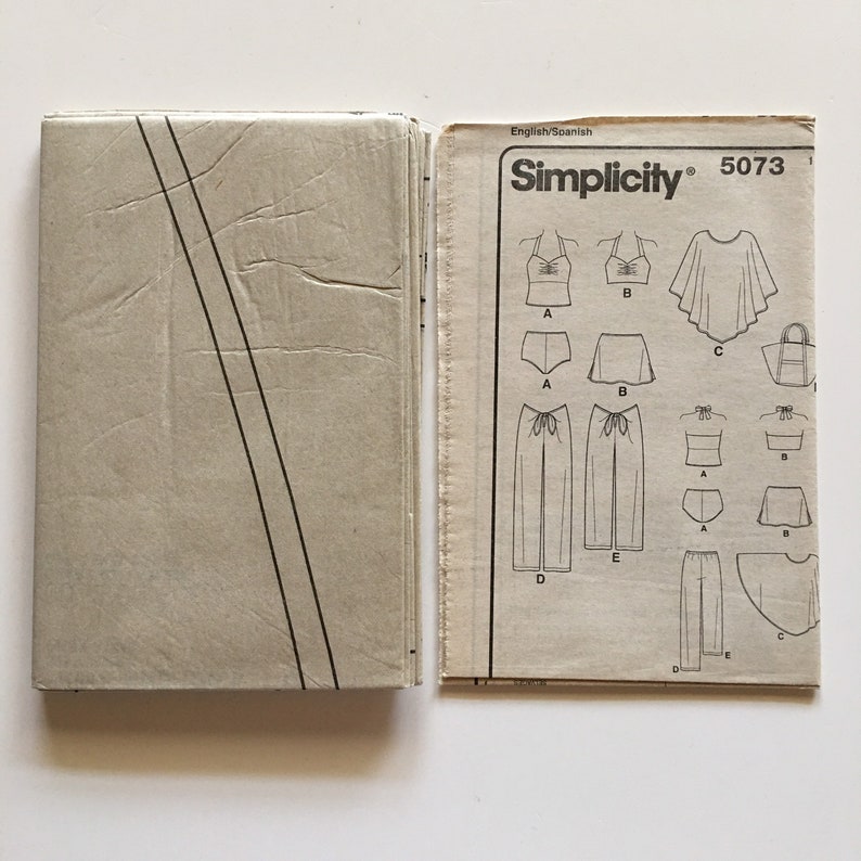 Puede incluir: Un patr&oacute;n de costura Simplicity vintage, modelo 5073, con ilustraciones de varias prendas de vestir, incluyendo tops, shorts, pantalones y un bolso tote. El patr&oacute;n est&aacute; en ingl&eacute;s y espa&ntilde;ol, con una combinaci&oacute;n de colores beige y blanco.