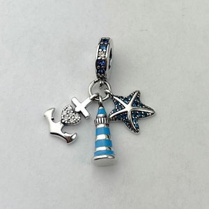 Könnte beinhalten: Ein silberner Charm mit maritimen Anhängern. Der Charm enthält einen Anker, ein Kreuz mit einem Herz, einen Leuchtturm und einen Seestern. Der Seestern und die Oberseite des Charms sind mit blauen Edelsteinen verziert.