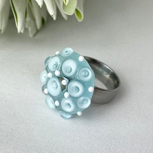 Puede incluir: Un anillo azul claro con un grupo de cuentas de vidrio blancas y azul claro. El anillo está hecho de acero inoxidable.