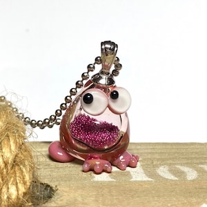 Peut inclure: Collier avec pendentif grenouille en verre rose, chaîne argentée et paillettes roses à l'intérieur. La grenouille a de grands yeux blancs avec des pupilles noires.