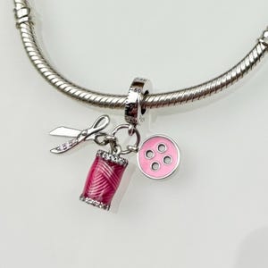 Op de afbeelding: Een zilveren bedelarmband met een naai-thema bedel. De bedel heeft een roze garenklos, een schaar en een roze knoop. De bedel is gedetailleerd met kleine, sprankelende accenten.