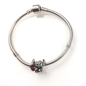 May include: A silver-toned snake chain charm bracelet featuring a single metal charm of a blue alien character holding a red flower and a green leaf. The bracelet includes a cylindrical clasp mechanism and two fixed stations along the chain.