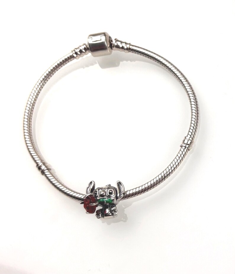 May include: A silver-toned snake chain charm bracelet featuring a single metal charm of a blue alien character holding a red flower and a green leaf. The bracelet includes a cylindrical clasp mechanism and two fixed stations along the chain.