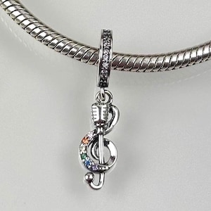 Op de afbeelding: Een zilveren bedel met een ontwerp van een G-sleutel met een regenboog van kleurrijke edelstenen. De bedel bevat ook een microfoon en hangt aan een zilveren kralenarmband.