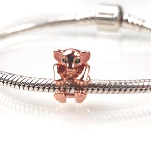 Könnte beinhalten: Ein roségoldfarbener Metall-Charm in Form eines kleinen Koalabären mit rosa emaillierten Ohren und dunklen Augen, befestigt an einem silberfarbenen Schlangenkettarmband mit einem zylindrischen Verschlussmechanismus am oberen Rand.