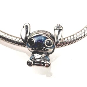 Puede incluir: Un charm de plata con la forma del personaje Stitch, con detalles en esmalte azul en las orejas, los ojos y la nariz. El charm est&aacute; insertado en una pulsera de cadena de serpiente de plata, mostrando un acabado met&aacute;lico pulido y rasgos esculpidos.