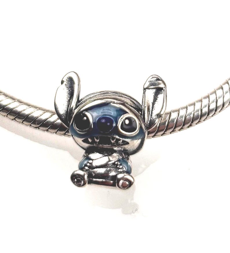 Puede incluir: Un charm de plata con la forma del personaje Stitch, con detalles en esmalte azul en las orejas, los ojos y la nariz. El charm est&aacute; insertado en una pulsera de cadena de serpiente de plata, mostrando un acabado met&aacute;lico pulido y rasgos esculpidos.