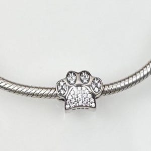 Op de afbeelding: Een zilverkleurige bedel in de vorm van een pootafdruk, bezet met kleine, sprankelende, heldere kristallen. De bedel is bevestigd aan een zilveren, getextureerde armband. Het pootafdrukontwerp is een populair symbool voor dierenliefhebbers.