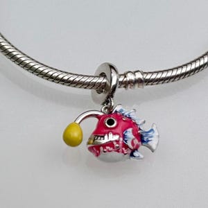 Pode incluir: Um charm colorido de peixe-pescador com acabamento em metal prateado. O peixe apresenta um corpo vermelho, barriga branca, barbatanas azuis e uma isca amarela. Este charm foi projetado para ser adicionado a uma pulseira.