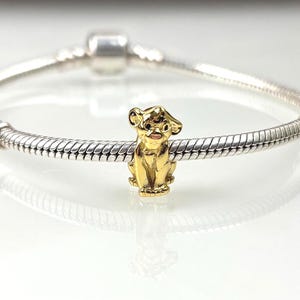 Könnte beinhalten: Ein goldfarbener Anhänger in Form eines Löwenjungen, der aufrecht sitzt. Der Anhänger ist an einem silbernen Armband befestigt. Das Löwenjunge hat detaillierte Merkmale, einschließlich Ohren, Nase und kleinem Mund. Der Anhänger ist wahrscheinlich aus Metall.