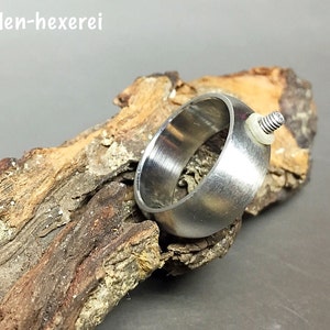 Könnte beinhalten: Ein silberner Ring mit einem Schraubendetail an der Seite. Der Ring liegt auf einem Stück Treibholz.