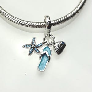 Op de afbeelding: Een zilveren bedelarmband met een verzameling bedels met oceaanthema. De bedels zijn onder meer een zeester met blauwe accenten, een teenslipper en een zeeschelp. De teenslipper en de zeeschelp zijn lichtblauw. De armband staat op een witte achtergrond.