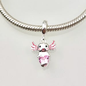 Può includere: Un charm d'argento con un design di axolotl a cartoni animati. Il charm ha dettagli rosa sulle branchie e un corpo di pietra preziosa rosa. Il charm è sospeso a una catena d'argento, perfetto per un braccialetto o una collana.