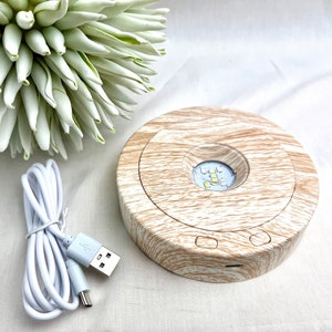 Könnte beinhalten: Ein Holz-Präsentationsständer mit einer runden Lichtbasis. Der Ständer hat ein helles Holzmaserungsmuster und ein weißes USB-Ladekabel.