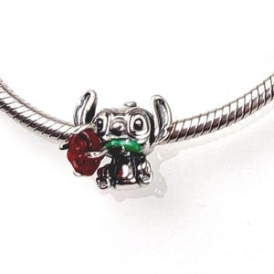 May include: A silver charm bead featuring a character with large ears holding a red rose with a green stem. The charm is threaded onto a silver snake chain bracelet, showcasing detailed metallic sculpting and vibrant red and green enamel accents.