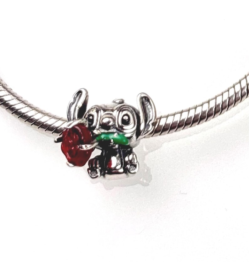 May include: A silver charm bead featuring a character with large ears holding a red rose with a green stem. The charm is threaded onto a silver snake chain bracelet, showcasing detailed metallic sculpting and vibrant red and green enamel accents.