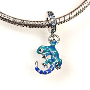 Könnte beinhalten: Ein silberner Anhänger in Form eines Geckos mit blau-türkiser Emaille-Verzierung, einem kleinen Auge und einem gestreiften Schwanz. Der Anhänger ist an einem Silberarmband befestigt und die Öse ist mit kleinen, klaren, funkelnden Steinen besetzt.