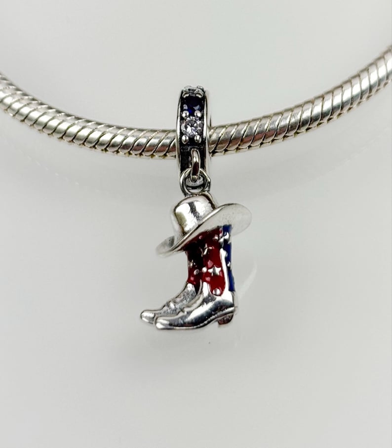 Pu&ograve; includere: Ciondolo in argento con un design di stivale e cappello da cowboy. Lo stivale &egrave; rosso, bianco e blu con stelle. Il ciondolo ha un cristallo trasparente nella parte superiore.
