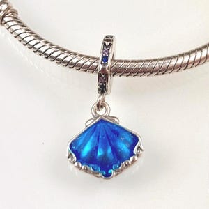 Pode incluir: Um berloque de pulseira em prata com um pingente de concha pendente com um acabamento em esmalte azul vibrante. A argola do pingente é decorada com pequenas pedras alternadas roxas e azuis cravadas numa estrutura de prata polida.