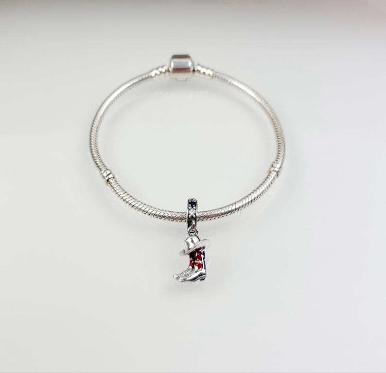 Pu&ograve; includere: Un braccialetto con ciondolo in argento con un ciondolo in argento a forma di stivale da cowboy con un design rosso, bianco e blu.