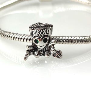 Könnte beinhalten: Ein silberner Charm-Anhänger mit einer stilisierten Figur mit baumartigem Kopf und grünen Augen. Der Charm ist mit geätzten Mustern versehen und hält kleine Objekte. Er wird an einem Silberarmband präsentiert.