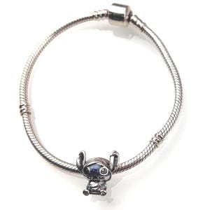 Puede incluir: Una pulsera de cadena de serpiente plateada con un dije de metal del personaje Stitch. El dije est&aacute; detallado con acentos de esmalte azul en las orejas y los ojos, y la pulsera incluye un cierre cil&iacute;ndrico en la parte superior.