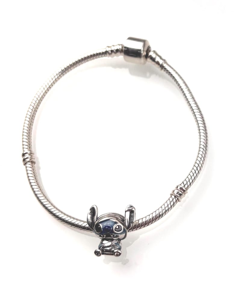 Puede incluir: Una pulsera de cadena de serpiente plateada con un dije de metal del personaje Stitch. El dije est&aacute; detallado con acentos de esmalte azul en las orejas y los ojos, y la pulsera incluye un cierre cil&iacute;ndrico en la parte superior.