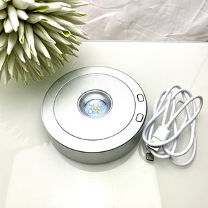 Op de afbeelding: Een ronde zilveren LED-lichtbasis met een USB-kabel. De lichtbasis heeft een kleine ronde opening in het midden met een wit LED-licht.