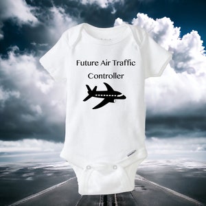 May include: White baby onesie with the text "Future Air Traffic Controller" and a black airplane graphic. The onesie is made of soft material and has short sleeves. The background features a cloudy sky and a runway.