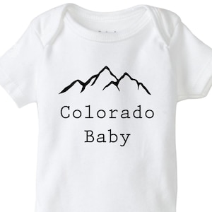 K&ouml;nnte beinhalten: Wei&szlig;er Baby-Strampler mit einem schwarzen Bergpanorama und dem Text "Colorado Baby".