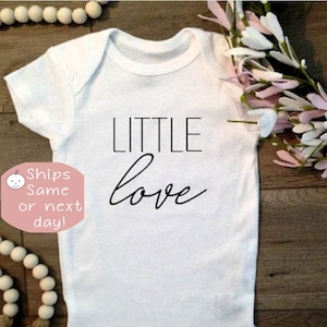 May include: White baby onesie with black text that reads "LITTLE love".