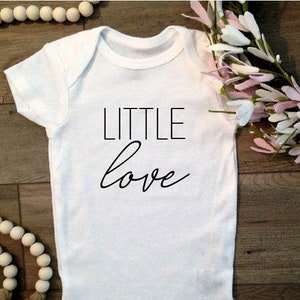 May include: White baby onesie with black text that reads "LITTLE love".