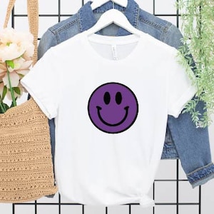 May include: White t-shirt with a purple smiley face graphic.