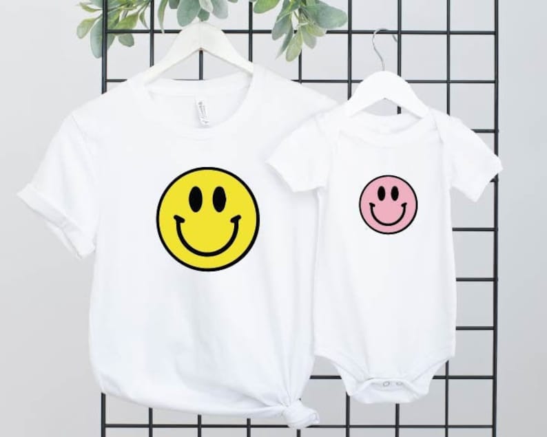 May include: A set of two white t-shirts featuring a smiley face design. The larger shirt has a yellow smiley face, while the smaller shirt has a pink smiley face.
