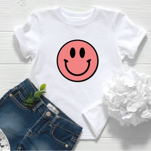 May include: A white t-shirt featuring a pink smiley face design. The shirt is styled with denim shorts and a white flower.
