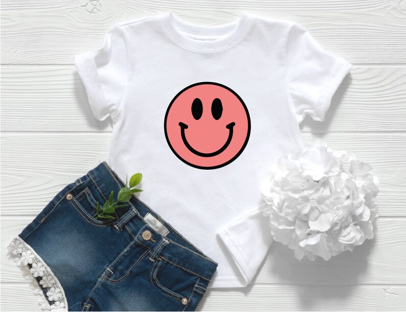 May include: A white t-shirt featuring a pink smiley face design. The shirt is styled with denim shorts and a white flower.