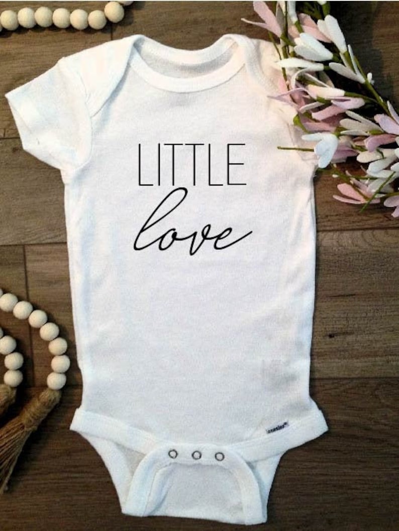 May include: White baby onesie with black text that reads "LITTLE love".