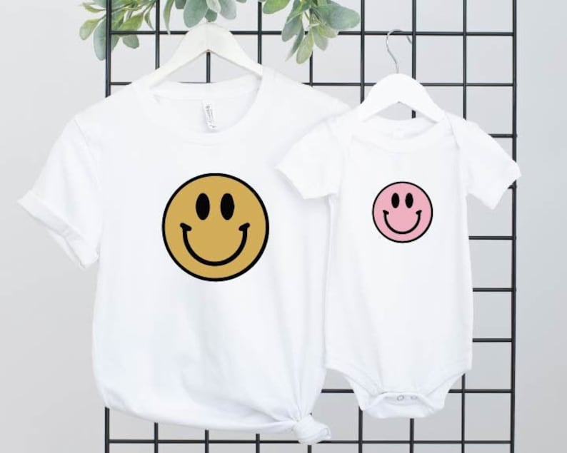 May include: Two white t-shirts with smiley faces. The larger shirt has a yellow smiley face with a black outline. The smaller shirt has a pink smiley face with a black outline.