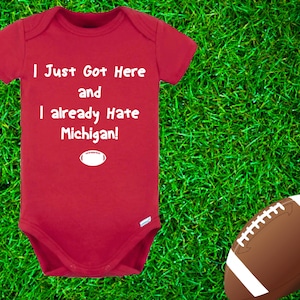 May include: Red baby onesie with white text that reads "I Just Got Here and I already Hate Michigan!" with a white football graphic below the text. The onesie is on a green grassy background with a brown football to the right.