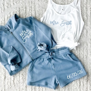 May include: A matching set of loungewear in light blue, including a zip-up hoodie, shorts, and a tank top. The hoodie and tank top feature the text "Mrs. Smith" in script, while the shorts display the date "06/25/26".