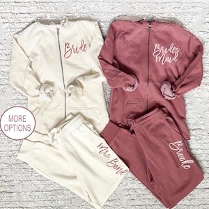 May include: Two sets of matching cream and maroon hooded sweatshirts and sweatpants. The cream set has the word "Bride" on the sweatshirt and "Mrs. Boyd" on the sweatpants. The maroon set has the words "Bridesmaid" on the sweatshirt and "Brooke" on the sweatpants.